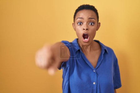 Photo for African american woman standing over yellow background pointing with finger surprised ahead, open mouth amazed expression, something on the front - Royalty Free Image
