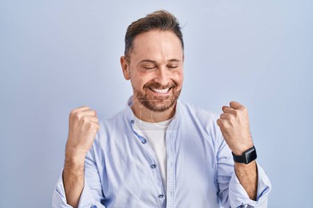 Téléchargez les photos : Middle age caucasian man standing over blue background very happy and excited doing winner gesture with arms raised, smiling and screaming for success. celebration concept. - en image libre de droit