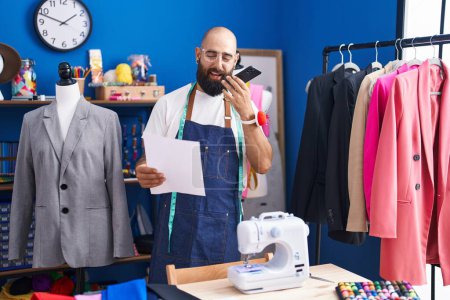 Photo for Young bald man tailor talking on smartphone looking clothing design at clothing factory - Royalty Free Image