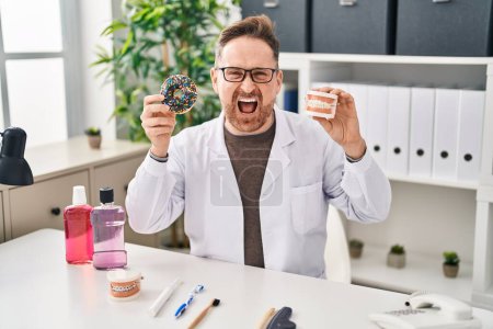 Photo for Middle age caucasian dentist man holding denture and doughnuts angry and mad screaming frustrated and furious, shouting with anger. rage and aggressive concept. - Royalty Free Image
