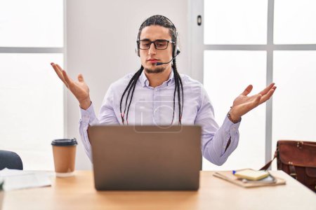 Téléchargez les photos : Homme hispanique aux cheveux longs travaillant à l'aide d'un ordinateur portable criant et criant fort à côté avec la main sur la bouche. concept de communication. - en image libre de droit