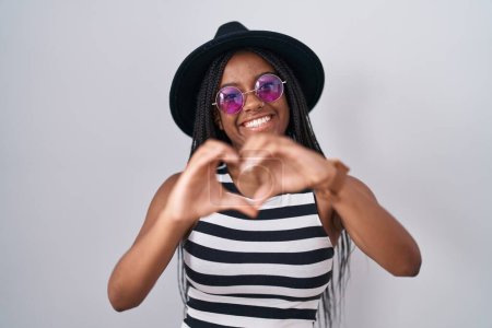 Photo for Young african american with braids wearing hat and sunglasses smiling in love doing heart symbol shape with hands. romantic concept. - Royalty Free Image
