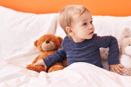 Foto de Adorable niño rubio sentado en la cama con oso de peluche en el dormitorio - Imagen libre de derechos