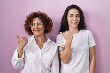 Foto de Madre e hija hispanas vistiendo una camiseta blanca casual sobre fondo rosa sonriendo con la cara feliz mirando y señalando hacia un lado con el pulgar hacia arriba. - Imagen libre de derechos