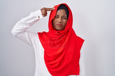 Téléchargez les photos : Jeune femme arabe portant une écharpe traditionnelle islamique en hijab tirant et se suicidant pointant la main et les doigts vers la tête comme une arme à feu, geste de suicide. - en image libre de droit