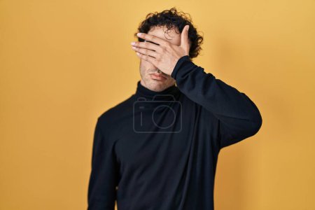Téléchargez les photos : Homme hispanique debout sur fond jaune couvrant les yeux avec la main, l'air grave et triste. concept de non-visibilité, de dissimulation et de rejet - en image libre de droit