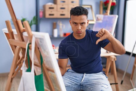 Foto de Joven hombre hispano pintando sentado en el estudio de arte con la cara enojada, signo negativo que muestra aversión con los pulgares hacia abajo, concepto de rechazo - Imagen libre de derechos