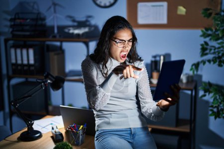 Photo for Young brazilian woman using touchpad at night working at the office pointing displeased and frustrated to the camera, angry and furious with you - Royalty Free Image