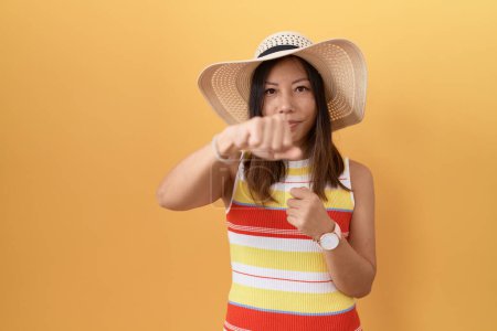 Photo for Middle age chinese woman wearing summer hat over yellow background punching fist to fight, aggressive and angry attack, threat and violence - Royalty Free Image