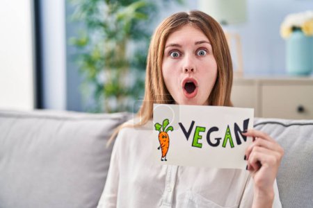 Foto de Beautiful woman holding banner with vegan word scared and amazed with open mouth for surprise, disbelief face - Imagen libre de derechos