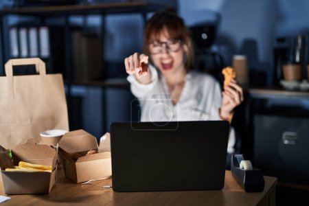 Photo for Young beautiful woman working using computer laptop and eating delivery food pointing displeased and frustrated to the camera, angry and furious with you - Royalty Free Image