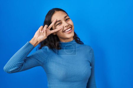 Photo for Young brazilian woman standing over blue isolated background doing peace symbol with fingers over face, smiling cheerful showing victory - Royalty Free Image