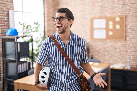 Téléchargez les photos : Homme hispanique aux cheveux longs travaillant au bureau tenant un casque de vélo en colère et fou hurlant frustré et furieux, criant de colère. rage et concept agressif. - en image libre de droit