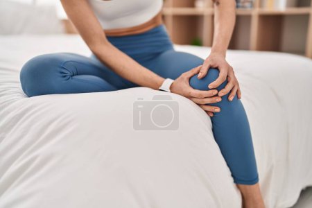 Foto de Young beautiful hispanic woman suffering for knee injury sitting on bed at bedroom - Imagen libre de derechos