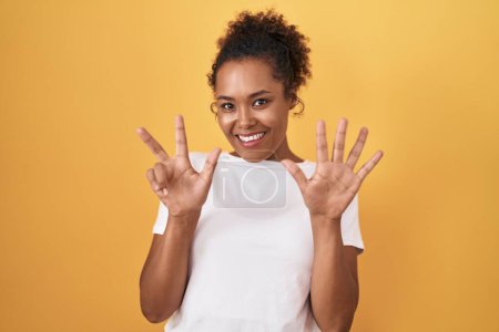 Photo for Young hispanic woman with curly hair standing over yellow background showing and pointing up with fingers number eight while smiling confident and happy. - Royalty Free Image