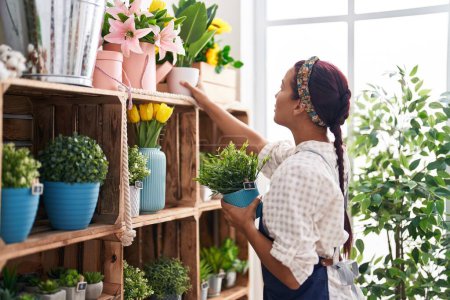 Téléchargez les photos : Jeune belle hispanique femme fleuriste tenant plante sur les étagères chez le fleuriste - en image libre de droit