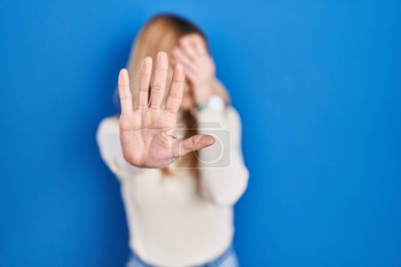 Téléchargez les photos : Jeune femme caucasienne debout sur fond bleu couvrant les yeux des mains et faisant arrêter geste avec expression triste et peur. concept embarrassé et négatif. - en image libre de droit