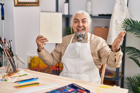 Photo for Middle age man with grey hair sitting at art studio holding notebook celebrating achievement with happy smile and winner expression with raised hand - Royalty Free Image