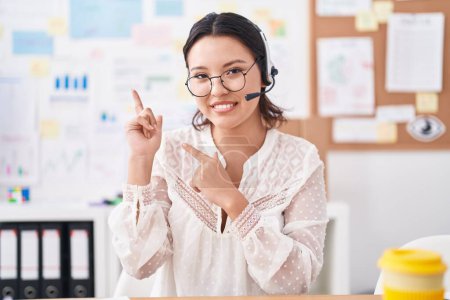 Téléchargez les photos : Hispanic young woman working at the office wearing headset and glasses smiling and looking at the camera pointing with two hands and fingers to the side. - en image libre de droit