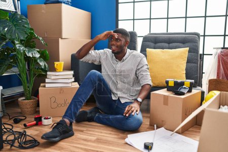 Téléchargez les photos : Homme afro-américain assis sur le sol à la nouvelle maison très heureux et souriant regardant loin avec la main au-dessus de la tête. concept de recherche. - en image libre de droit
