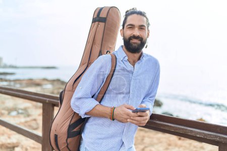 Foto de Young hispanic man musician using smartphone holding guitar case at seaside - Imagen libre de derechos