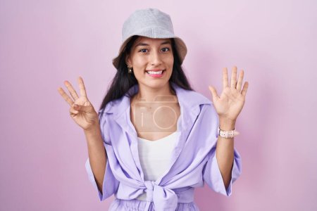 Foto de Young hispanic woman standing over pink background wearing hat showing and pointing up with fingers number eight while smiling confident and happy. - Imagen libre de derechos