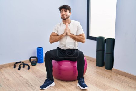 Téléchargez les photos : Hispanic man with beard sitting on pilate balls at yoga room begging and praying with hands together with hope expression on face very emotional and worried. begging. - en image libre de droit