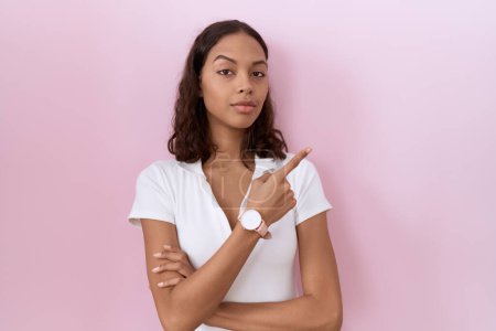 Foto de Mujer hispana joven con camiseta blanca casual apuntando con el dedo de la mano hacia un lado mostrando publicidad, cara seria y tranquila - Imagen libre de derechos