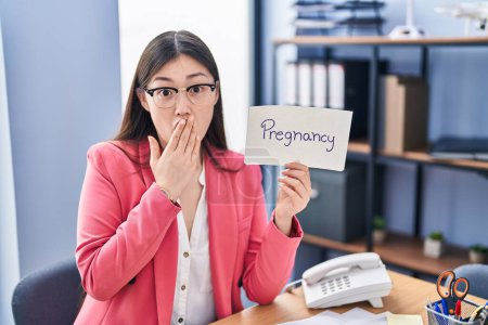Foto de Chinese young woman working at the office holding pregnancy sign covering mouth with hand, shocked and afraid for mistake. surprised expression - Imagen libre de derechos