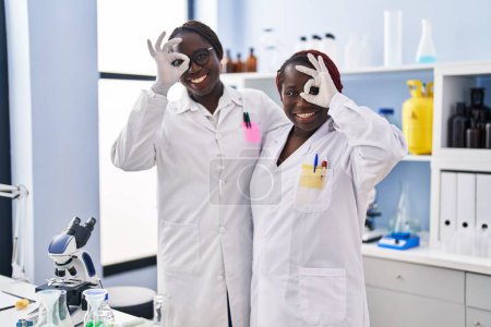 Foto de Dos mujeres africanas que trabajan en el laboratorio científico sonriendo felices haciendo bien signo con la mano en el ojo mirando a través de los dedos - Imagen libre de derechos