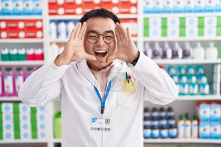 Foto de Chino joven que trabaja en farmacia droguería sonriendo alegre jugando a echar un vistazo a un boo con las manos mostrando la cara. sorprendido y salido - Imagen libre de derechos