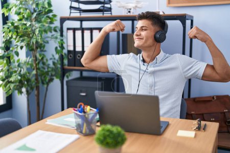 Foto de Joven hombre hispano trabajando en la oficina con auriculares que muestran los músculos de los brazos sonriendo orgulloso. concepto de fitness. - Imagen libre de derechos