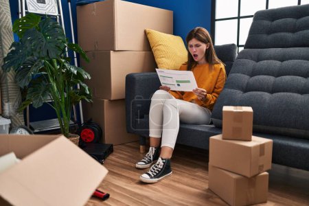 Foto de Young brunette woman sitting on the sofa at new home looking at documents scared and amazed with open mouth for surprise, disbelief face - Imagen libre de derechos