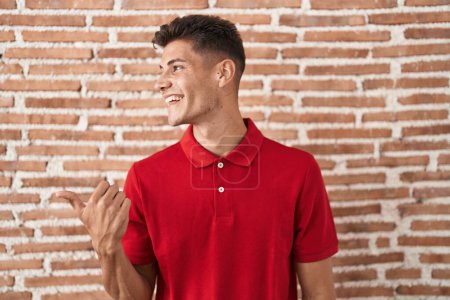 Foto de Joven hombre hispano de pie sobre la pared de ladrillos sonriendo con la cara feliz mirando y señalando a un lado con el pulgar hacia arriba. - Imagen libre de derechos