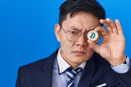 Foto de Joven hombre asiático sosteniendo bitcoin moneda virtual que cubre ojo despistado y expresión confusa. concepto de duda. - Imagen libre de derechos