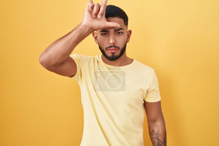 Téléchargez les photos : Jeune homme hispanique debout sur fond jaune se moquant des gens avec les doigts sur le front faisant geste de perdant se moquant et insultant. - en image libre de droit