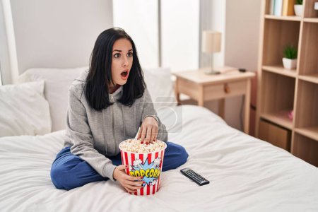 Foto de Mujer hispana comiendo palomitas de maíz viendo una película en la cama con cara de shock, mirando escéptica y sarcástica, sorprendida con la boca abierta - Imagen libre de derechos