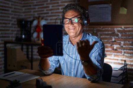 Foto de Hombre hispano de mediana edad usando touchpad sentado en la mesa por la noche apuntando hacia atrás con la mano y los pulgares hacia arriba, sonriendo confiado - Imagen libre de derechos