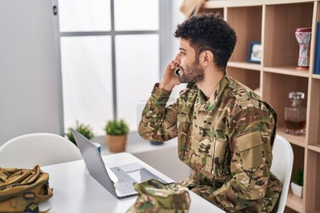 Téléchargez les photos : Young arab man army soldier using laptop talking on the smartphone at home - en image libre de droit