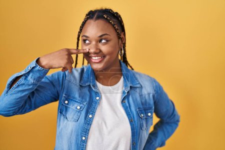 Téléchargez les photos : Femme afro-américaine avec des tresses debout sur fond jaune pointant du doigt vers le visage et le nez, souriante et joyeuse. concept de beauté - en image libre de droit