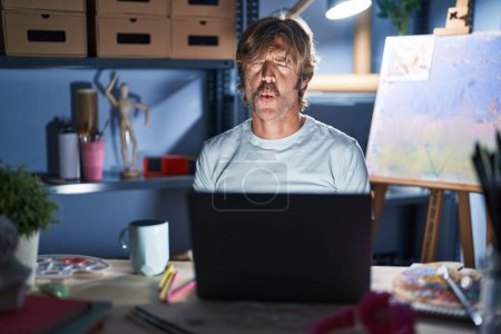 Photo for Middle age man sitting at art studio with laptop at night making fish face with lips, crazy and comical gesture. funny expression. - Royalty Free Image