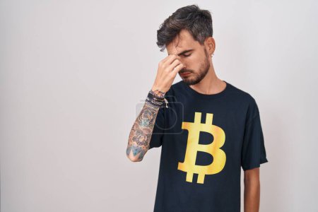 Foto de Young hispanic man with tattoos wearing bitcoin t shirt tired rubbing nose and eyes feeling fatigue and headache. stress and frustration concept. - Imagen libre de derechos