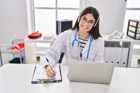 Young beautiful hispanic woman doctor having video call writing on document at clinic