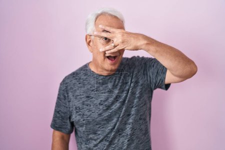 Photo for Middle age man with grey hair standing over pink background peeking in shock covering face and eyes with hand, looking through fingers with embarrassed expression. - Royalty Free Image