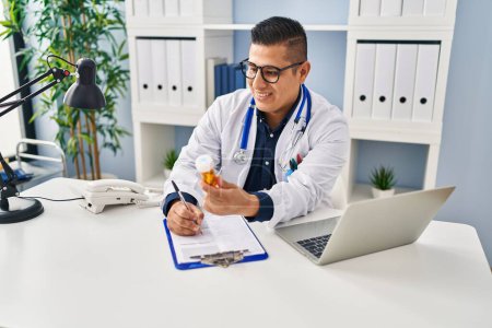 Photo for Young latin man doctor writing on document holding pills at clinic - Royalty Free Image