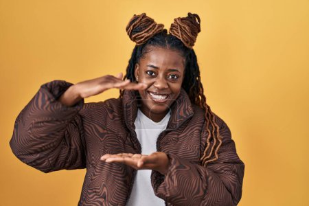Téléchargez les photos : Femme africaine avec les cheveux tressés debout sur fond jaune geste avec les mains montrant grand et grand signe de taille, symbole de mesure. souriant en regardant la caméra. concept de mesure. - en image libre de droit