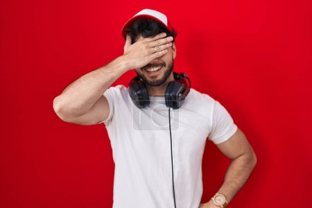 Téléchargez les photos : Hispanic man with beard wearing gamer hat and headphones smiling and laughing with hand on face covering eyes for surprise. blind concept. - en image libre de droit