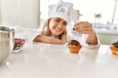 Téléchargez les photos : Young woman wearing cook uniform putting raspberry on cupcake at kitchen - en image libre de droit