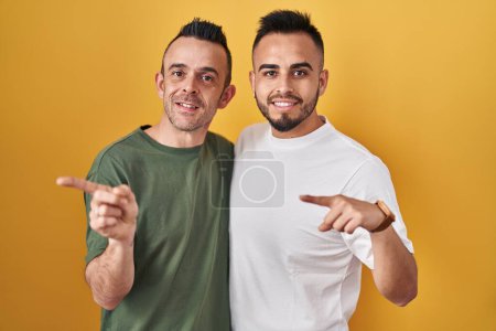 Téléchargez les photos : Couple homosexuel debout sur fond jaune pointant vers le dos derrière avec la main et les pouces levés, souriant confiant - en image libre de droit