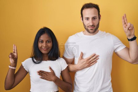 Téléchargez les photos : Couple Interracial debout sur fond jaune souriant jurer avec la main sur la poitrine et les doigts vers le haut, faisant un serment de fidélité promesse - en image libre de droit